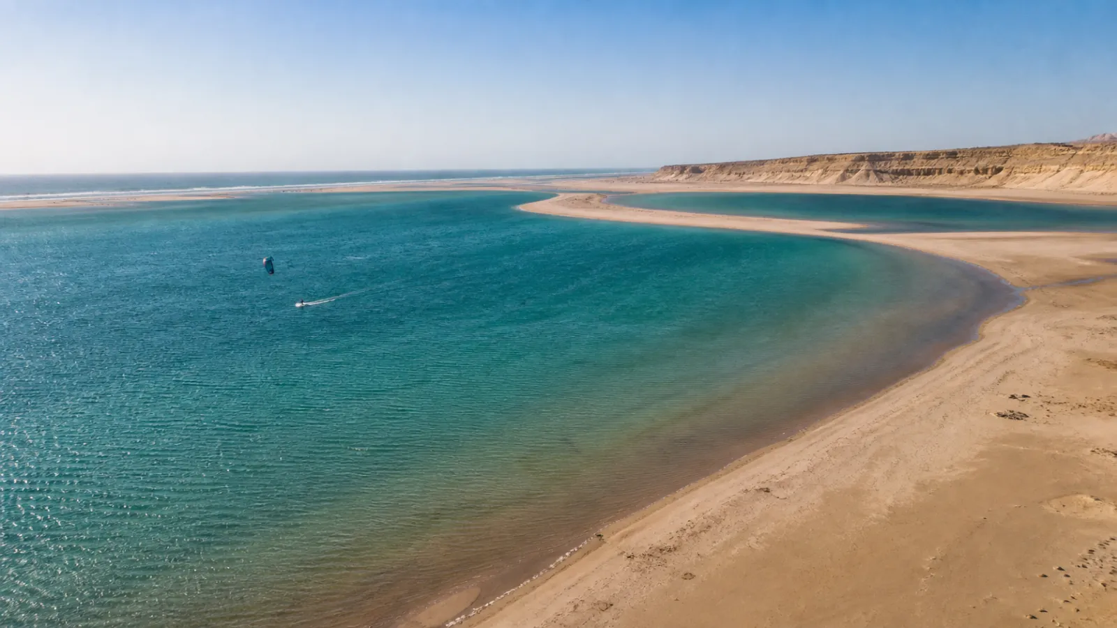 Turquoise Dakhla lagoon bordered by desert sand with a kitesurfer on the water