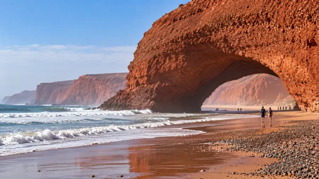 Travelers walking beneath the red rock arch at Legzira Beach on a coastal trip from Agadir