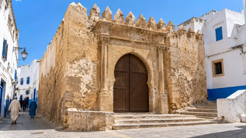 Bab Homar historic Medina gate in Asilah, Morocco, featuring weathered stone walls, a traditional wooden entrance and whitewashed buildings.