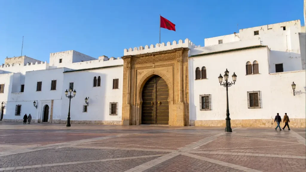 Royal Palace exterior at Place Hassan II in Tetouan, Morocco, featuring an ornate traditional gateway, white palace walls and the Moroccan flag.